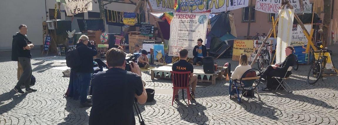 Ein Foto der Pressekonferenz für Pimmelgate Süd. Alex sitzt vor dem Klimacamp. Vor ihm sitzen im Halbkreis etwa ein dutzend Menschen um ihn herum. Kameras und Fotoapparate sind auf ihn gerichtet.