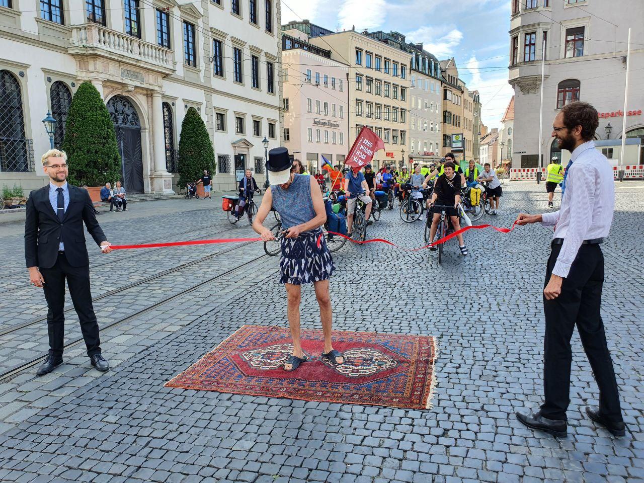 Am Rathausplatz vor dem Klimacamp halten zwei Klimaaktivist*innen ein rotes Band über einen dort ausgelegten Teppich, während ein* Dritte*r das Band gerade durchschnitten hat. Eine Hälfte des Bandes flattert bereits davon. Im Hintergrund warten über dreißig Radfahrer*innen darauf ihre Tour mit einer Fahrt über den Teppich zu beginnen.