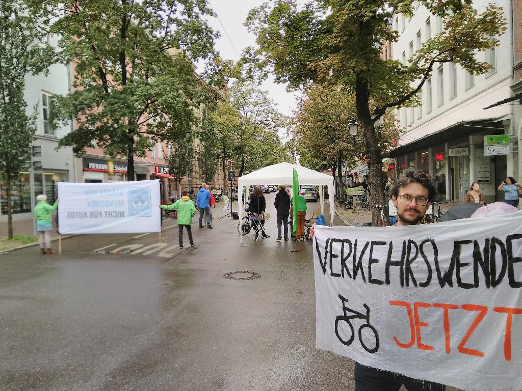 Das Bild zeigt die Bahnhofstraße. Rechts im Bild hält ein Verkehrswendeaktivist ein Plakat mit der Aufschrift „Verkehrswende jetzt“ in die Kamera. Links im Bild stehen zwei Aktivist*innen von Greenpeace, die ein großes Banner mit der Aufschrift „Städte für Menschen nicht für Autos“ in die entgegengesetzte Richtung halten. Der Schriftzug schimmert durch den Stoff des Banners durch. Weiter sieht man mehrere Aktivist*innen, Passant*innen, einen Pavillion, kleinere Klappstühle- und tische, Lautsprecher sowie im Hintergrund ein Polizeiauto. Gerade so nicht mehr im Bild erkennbar sind Aktivist*innen, die hinter dem Pavillion auf der Straße Federball spielen. Der Boden ist feucht, weil es kurz zuvor geregnet hatte. Links und rechts der Straße stehen Bäume, deren Blätterdach sich über die Straße spannt.