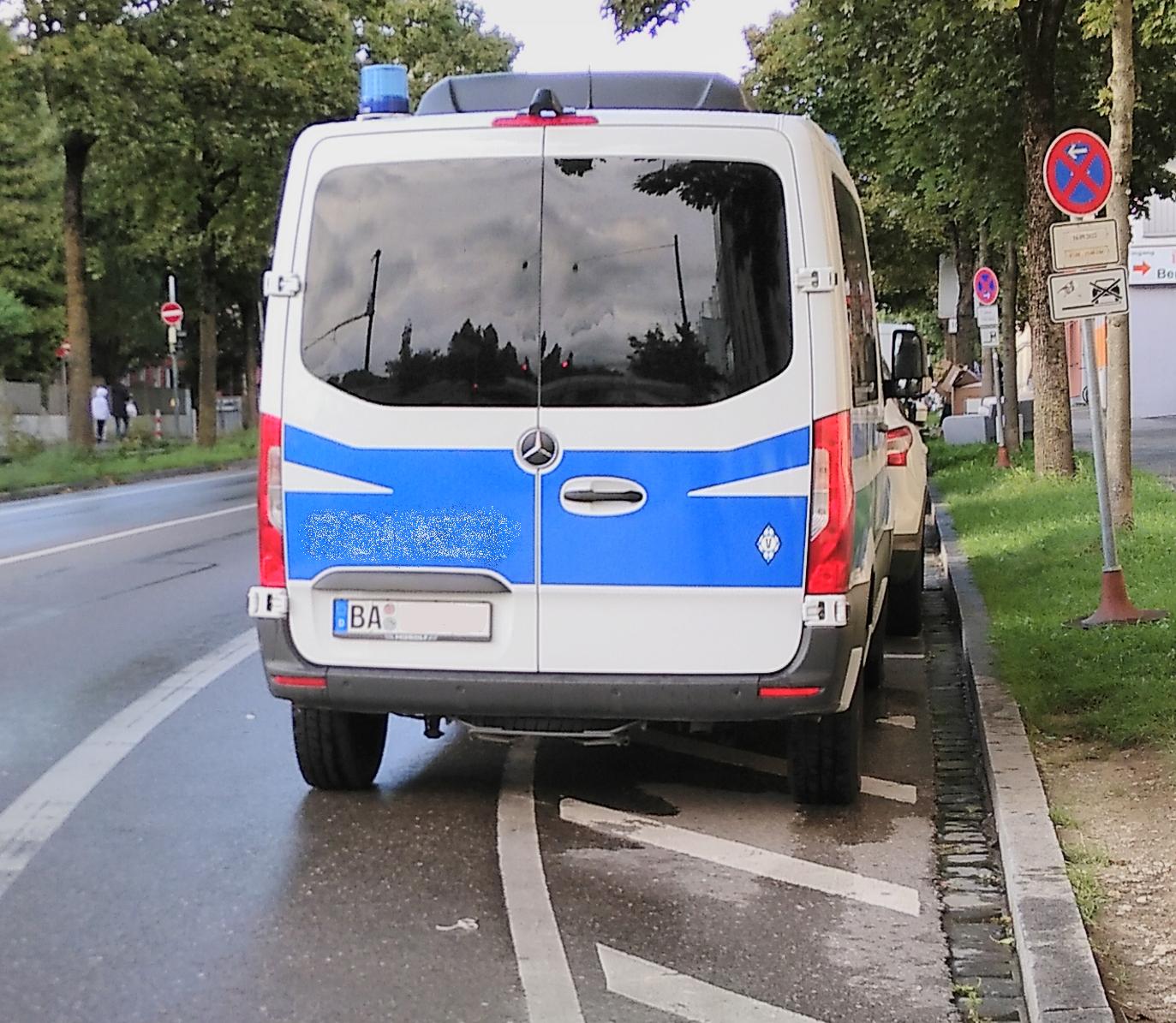 Das Foto zeigt einen Einsatzwagen der bayerischen Bereitschaftspolizei, der im Halteverbot steht und so weit in den Radweg hineinreicht, dass Fahrradfahrer*innen auf die Straße ausweichen müssen. Die Autonummer und der Schriftzug „Polizei“ wurden verpixelt.