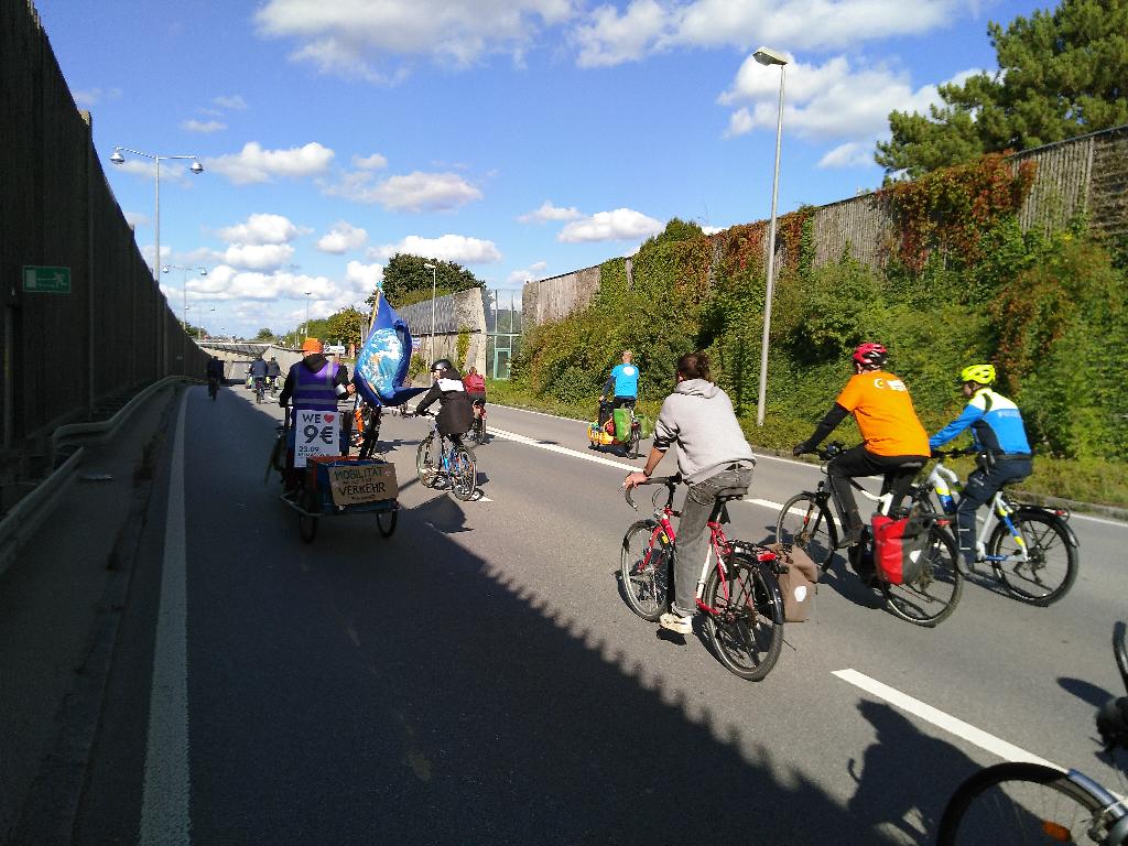 Das Bild zeigt Fahrradfahrer auf der B17. Der Asphalt ist grau, der Himmel ist blau gesprenkelt mit einzelnen weißen Wolken. Auf beiden Seiten der Straße sind Lärmschutzwände. Eine der Lärmschutzwände schirmt auch den Blick auf die Gegenfahrbahn ab. Die Radfahrer verteilen sich auf die zwei Fahrspuren. Zusätzlich gibt es noch eine Abbiegespur. In der Entfernung sieht man eine Brücke über die B17.