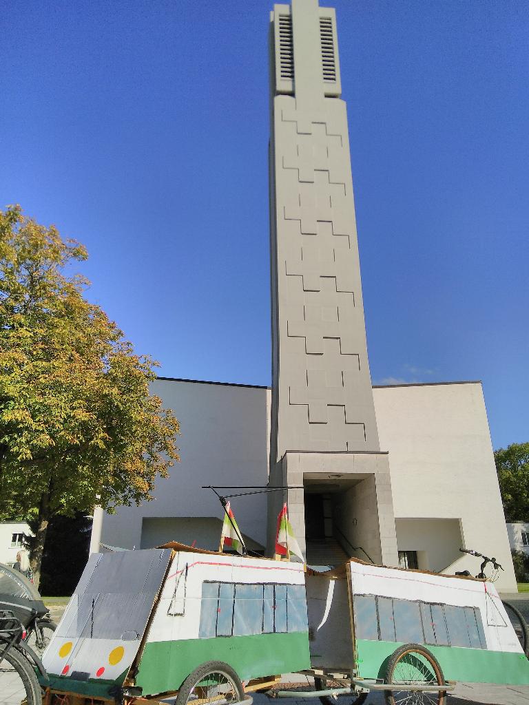 Das Bild zeigt die Modellstraßenbahn auf dem Zwölf-Apostel-Platz. Das Bild ist hochkant, damit im Hintergrund noch der Turm der Kirche „Zu den Heiligen Zwölf Aposteln“ drauf passt. 