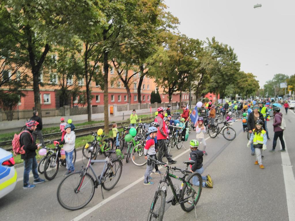 Das nördliche Ende der Haunstetter Straße ist voller herumtollender Kinder, herumgehender Erwachsener und abgestellter Fahrräder. Anb einigen Fahrrädern sind Luftballons oder Fahnen angebracht. Das Bild ist von der vorderen linken Ecke der Demo aufgenommen worden. Im Hintergrund sieht man die mit Bäumen flankierte Straßenbahnstrecke der Linien 2 und 3. Am linken Bildrand sieht man noch das Heck eines der beiden Polizeiautos, welche der Demo voran fuhren.