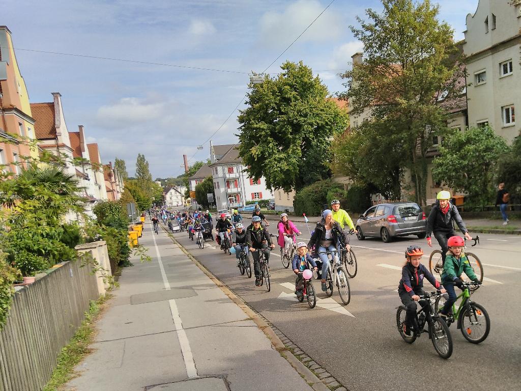 Das Foto ist vom Gehweg nahe der Einmündung der Hochfeldstraße in die Schertlinstraße aus aufgenommen. Von dort fällt die Schertlinstraße nach Osten hin ab. Das Foto zeigt einen langen Strom von Fahrradfahrer*innen, die wie sie die Schertlinstraße hinauf fahren. Viele der kleinsten Fahrradfahrer*innen fahren auf der rechten Seite der Straße nahe am Gehweg, während es vor allem erwachsene Fahrradfahrer*innen sind, die nahe des Gegenverkehrs fahren.