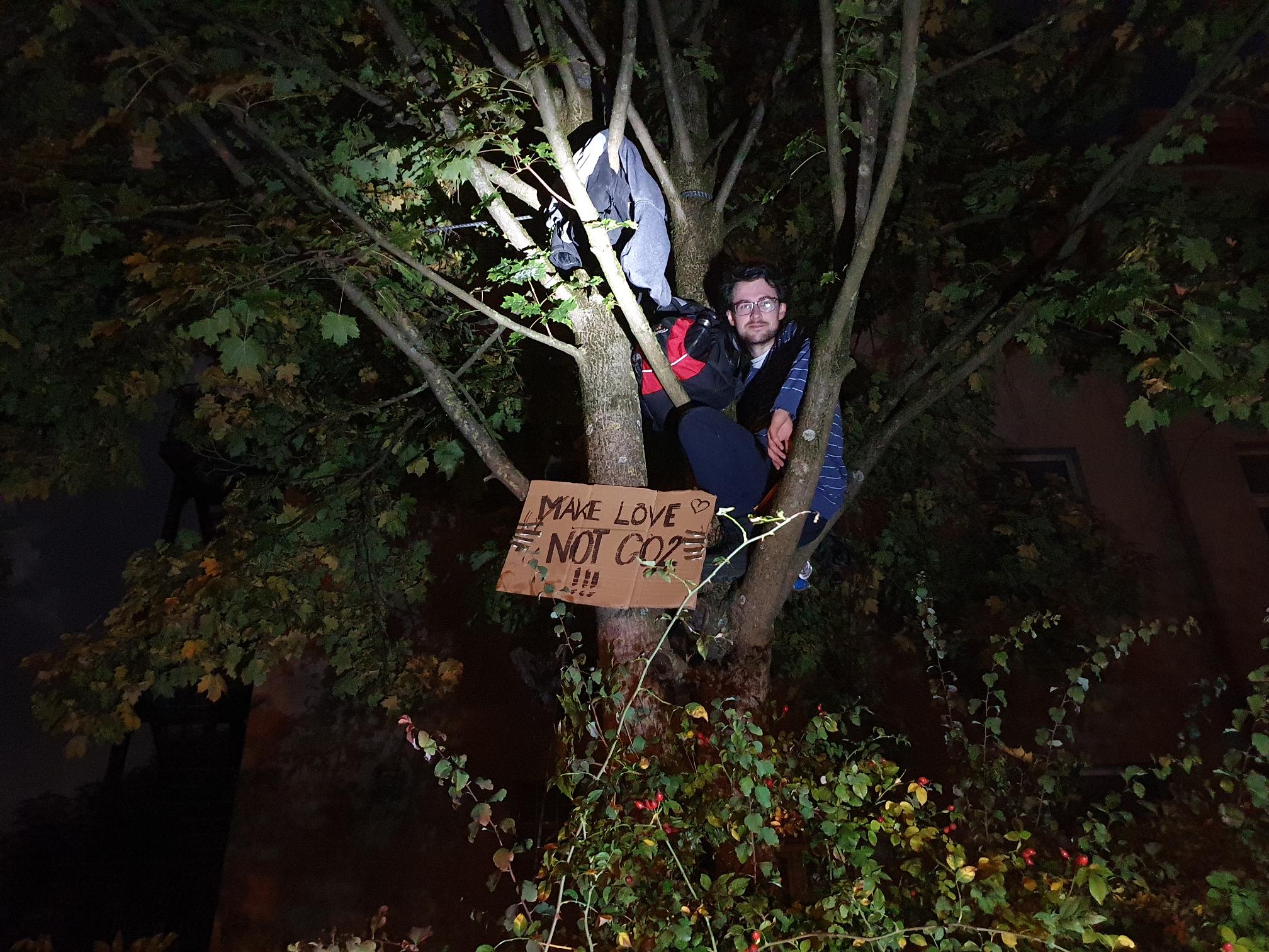 Ein Aktivist in einem Baum. Unterhalb von ihm hängt ein Pappschild mit der Aufschrift „Make Love not CO2!!!“. Das Bild ist in den frühen Morgenstunden noch im Dunkeln aufgenommen worden. Ein Scheinwerfer oder eine Lampe leuchten den Aktivisten an.