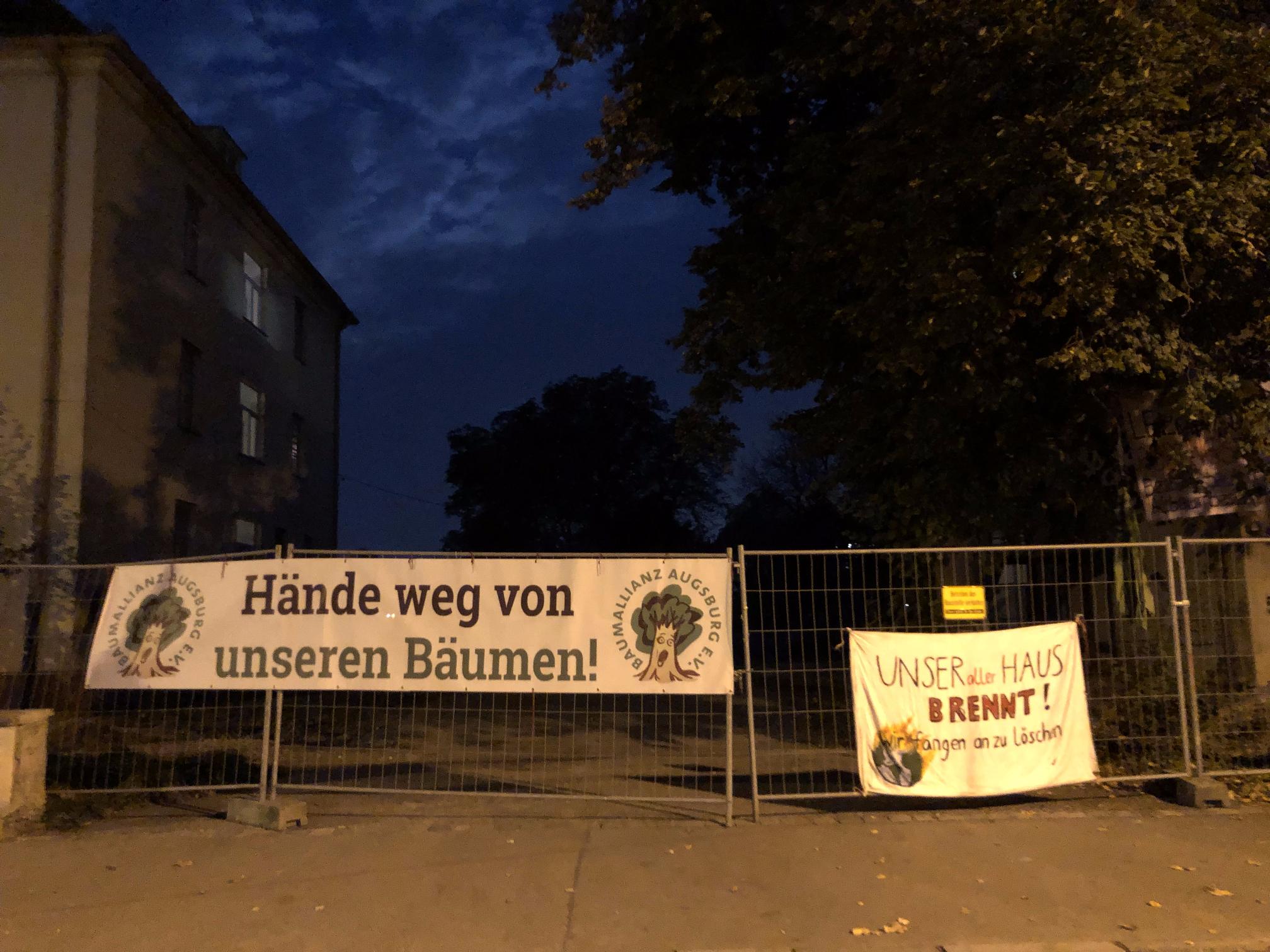 An einem Bauzaun vor dem Reese-Areal hängen zwei Banner. Auf dem linken Banner steht „Hände weg von unseren Bäumen“ und „BAUMALLIANZ AUGSBURG E.V.“. Es sind Bäume darauf abgebildet. Das rechte Banner zeigt eine brennende Erdkugel. Handgeschrieben steht darauf „UNSER aller HAUS BRENNT! Wir fangen an zu löschn“. Im Hintergrund sind ein altes Gebäude und mehrere schöne alte große Laubbäume zu sehen. Das Bild ist in den frühen Morgenstunden aufgenommen. Zwischen den Wolken erhellt sich langsam der Himmel.