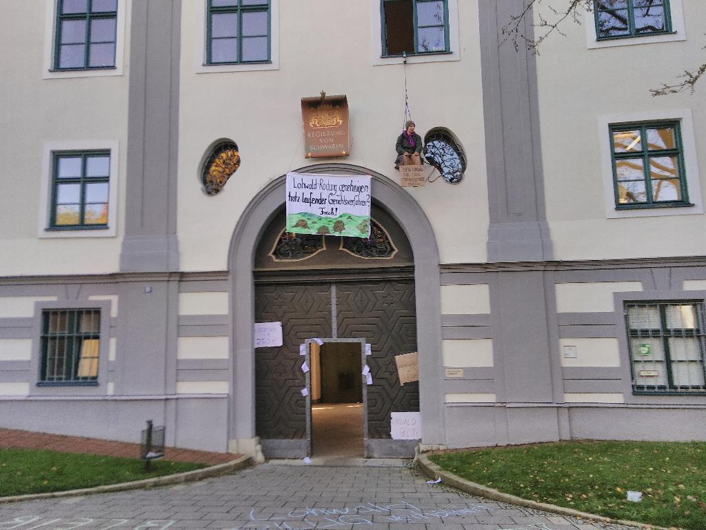 Das Bild zeigt den Eingang zum Regierungsgebäude. Über dem Eingang am Schild mit der Beschriftung „Regierung von Schwaben“ hängt in etwa 3 bis 4 Metern Höhe ein Banner mit der Aufschrift „Lohwald-Rodung genehmigen trotz laufender Gerichtsverfahren? Frech!“. Nochmal ein wenig höher rechts von der Tür sitzt eine Aktivistin an einem Seil hängend, welches aus einem Fenster des zweiten Stockes hängt.