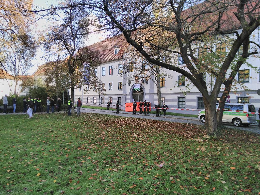 Links im Bild der Park des Fronhofes, in dem Passant*innen, Vertreter*innen der Presse und Polizist*innen für Kommunikation und Pressearbeit. Rechts im Bild befindet sich das Regierungsgebäude mit der am Seil sitzenden Aktivistin und neun in der Gegend herumstehenden Polizist*innen.