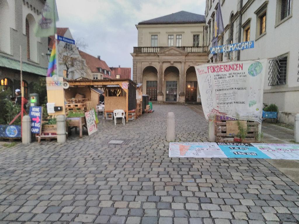 Das Bild zeigt das Klimacamp aus Richtung des Rathausplatzes. Links stehen einige Platzparks sowie Schilder mit politischen Aussagen. Dahinter befindet sich ein überdachter nach Vornen offener Aufenthaltsbereich. Dahinter befinden sich die übrigen funktionalen Aufbauten des Camps. In der Mitte ist eine Zufahrt zum Fischmarkt freigehalten worden. Auf der rechten Seite hängt das Banner mit den Forderungen des Camps. Davor liegt ein Bodenbanner mit Informationen über Wind- und Solarenergie. Das Foto enstand am Abend des 05.11.2022.