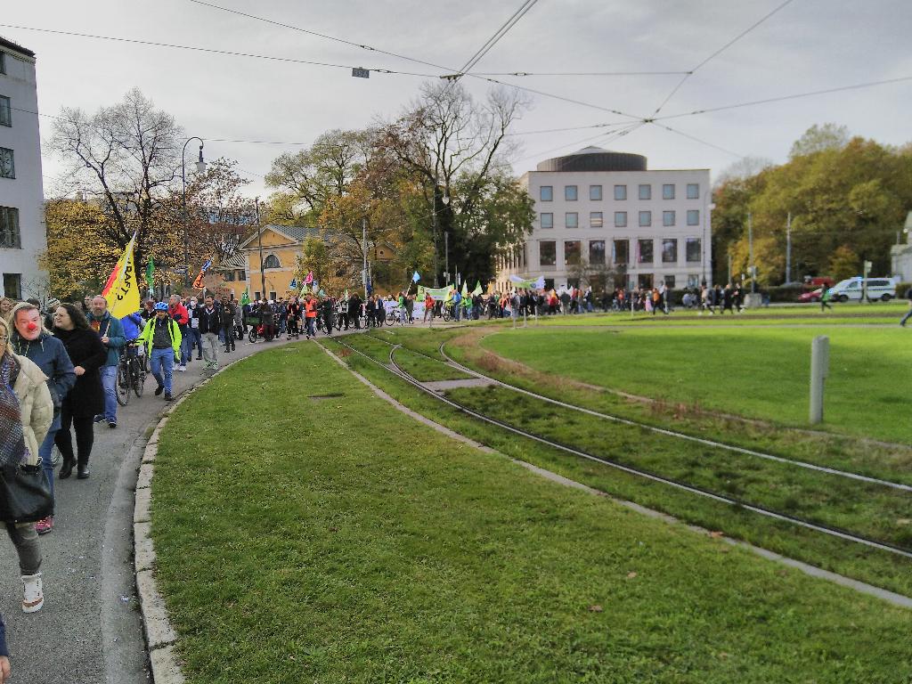 Der Karolinenplatz ist ein Kreisverkehr mit etwa hundert Metern Durchmesser. Das Bild wurde von Vornen im Demonstrationszug aufgenommen und zeigt den hinteren Teil der Demonstration, wie er gerade im Kreisverkehr unterwegs ist. Links im Bild trägt eine Person eine ‚Klimaclown Söder‘-Maske.