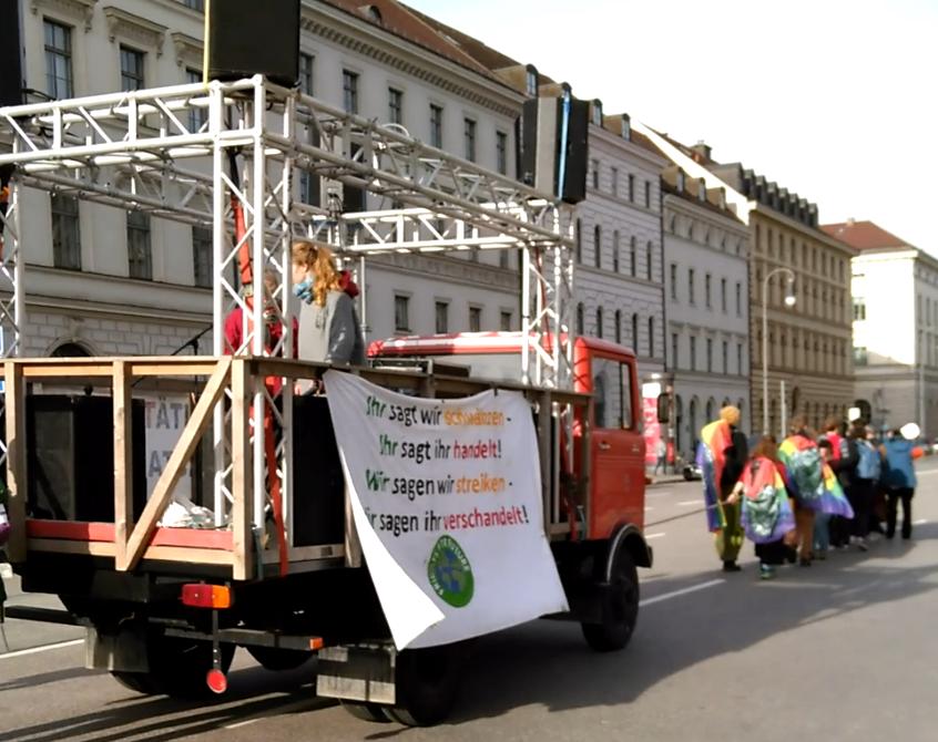 Der Lautsprecherwagen wird auf der Maximilianstraße von zehn oder mehr Demonstrationsteilnehmer*innen an einem dicken langen Seil gezogen. Der Lautsprecherwagen selbst ist ein umgebautes altes Feuerwehrauto, auf dessen Ladefläche ein Gerüst mit einem Lautsprecherturm in jeder Ecke und einem Geländer angebracht wurde. Auf der Ladefläche selbst stehen zwei junge Aktivistinnen.