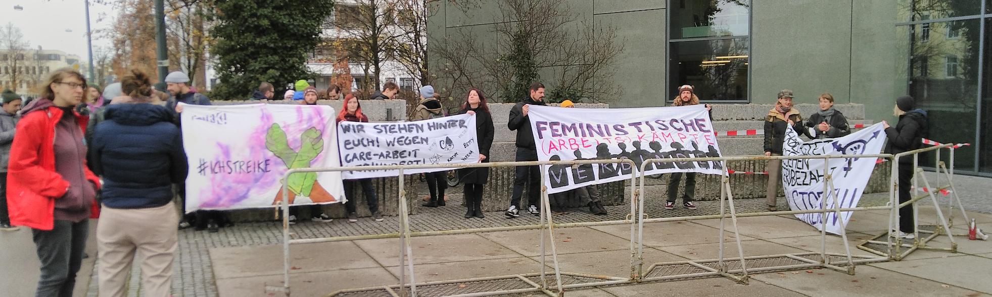 Das Bild zeigt eine Demo vor dem Gerichtssaal. Es sind vier Plakete zu erkennen. Darauf steht: „#ichStreike“, „Wir stehen hinter euch! Wegen Care-Arbeit verhindert!“ gefolgt von einer Liste von Namen, „Feministische (Arbeits-)Kämpfe verbinden“ und „Überbelastet Ungesehen Ungesehen Wir streiken!