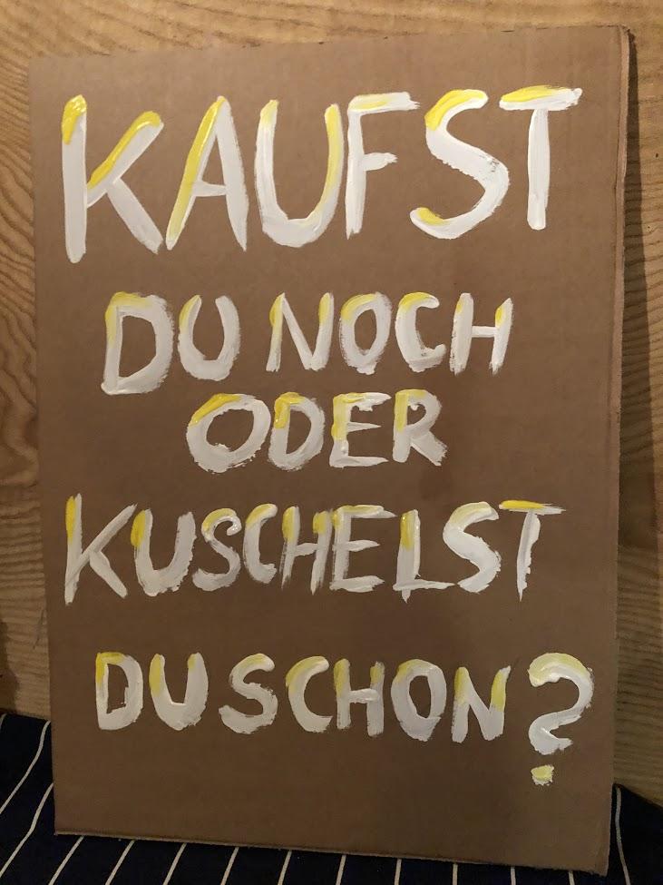 Das Bild zeigt ein Pappschild mit der Aufschrift „Kaufst du noch oder kuschelst du schon?“.