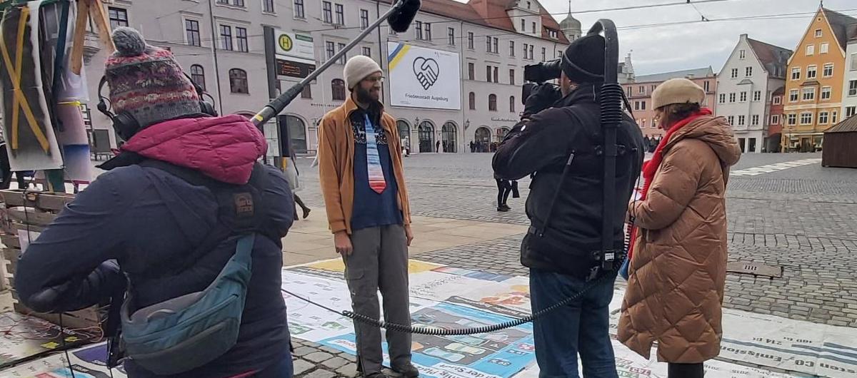 Vor dem Klimacamp wird ein Klimagerechtigkeitsaktivist von einem dreiköpfigen Fernsehteam interviewt. Eine Person hält ein Mikrofon, eine Person hält eine Kamera und die dritte Person liest von einem Notizzettel. Auf dem Boden liegen Bodenbanner, die über verschiedene Aspekte der Klimakrise informieren. Im Hintergrund der Rathausplatz und das große „Friedensstadt Augsburg“-Plakat.