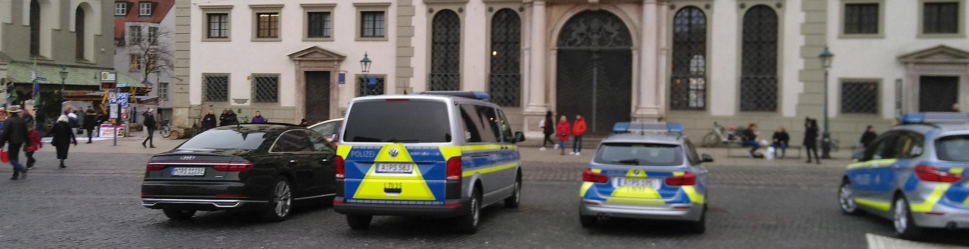 Vom Rathausplatz aus gesehen links das Klimcamp und rechts das Rathaus. Vor dem Klimacamp stehen einige Menschen in Gespräche vertieft. Es ist kaum zu erkennen, aber darunter befindet sich auch ein Kameraperson. Vor dem Rathaus stehen ein teures Auto, welches nach Politiker*in aussieht, und drei Polizeiautos.