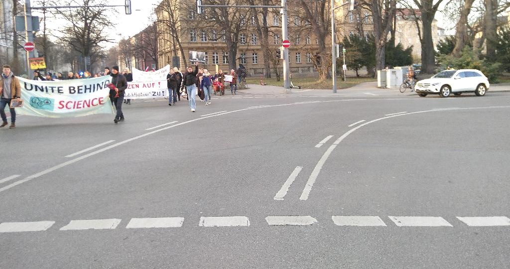 Das Bild zeigt links die Demonstrationsteilnehmer*innen, wie sie von Norden die Schaezlerstraße entlang auf die Kamera zulaufen, und rechts ein auf einer Querstraße, genannt „Am Alten Einlaß“, wartendes Auto. Zwischen Demo und Auto sind vielleicht fünfzehn Meter freie Asphaltfläche und keinerlei Polizei ist in Sichtweite. Auf einem großen von Demonstrationsteilnehmer*innen getragenen Plakat steht „UNITE BEHIND SCIENCE“. In Abstand dahinter gibt es ein paar kaum leserliche Tierschutzplakate mit Sprüchen wie „GO VEGAN OR DIE“, „Tiere sind keine Ware“ und unleserlich teilweise verdeckt irgendetwas mit „...zen auf!“ und „...Schlachthäuser ZU!“. Hinzu kommen weitere unleserliche Plakete in der Ferne.