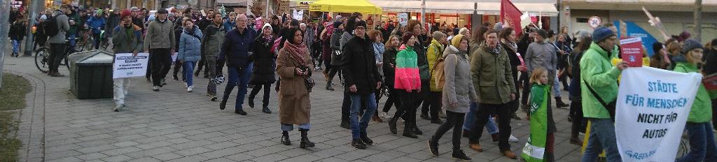 Das Foto zeigt einen Abschnitt aus der Mitte des Demonstrationszuges, wie er aus Richtungs Schaezlerstraße den Königsplatz betritt. Die Gruppe ist sehr heterogen mit Frauen und Männern von Kindesalter bis Rentenalter. Am rechten Rand tragen ein Mann und eine Frau von Greenpeace gemeinsam ein Plakat mit dem Spruch „Städte für Menschen nicht für Autos“. Dahinter etwas weiter links läuft ein junges Mädchen in eine ‚Fridays for Future‘-Flagge eingewickelt. Noch weiter hinten auf der linken Bildseite geht ein Mann, der am Handy telefoniert und ein Plakat mit einem unleserlichenVerweis auf das 9-Euro-Ticket trägt. Mehrere Menschen tragen Zettel mit der Aufschrift „KEINE NEUEN AUTOBAHNEN“ mit sich. Die Aufschriften auf weiteren Plakaten im Hintergrund sind nicht zu entziffern.