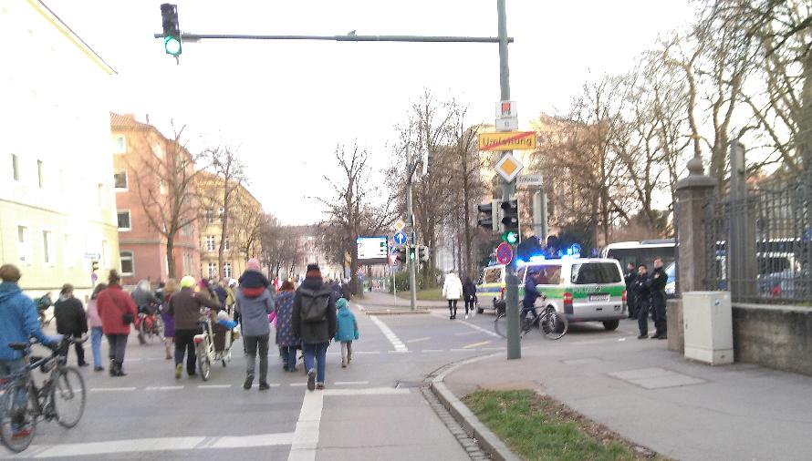 Auf der linken Seite sieht man die Demonstration die Schaezlerstraße entlang gehen. Es sind junge und ältere Menschen, viele Familien und mehrere mitgeschobene Fahrräder darunter. Rechts sieht man die Einmündung der Frölichstraße in die Schaezlerstraße. Die Einmündung wird durch drei Einsatzwägen blockiert. Zwischen den Einsatzwägen stehen Polizist*innen.