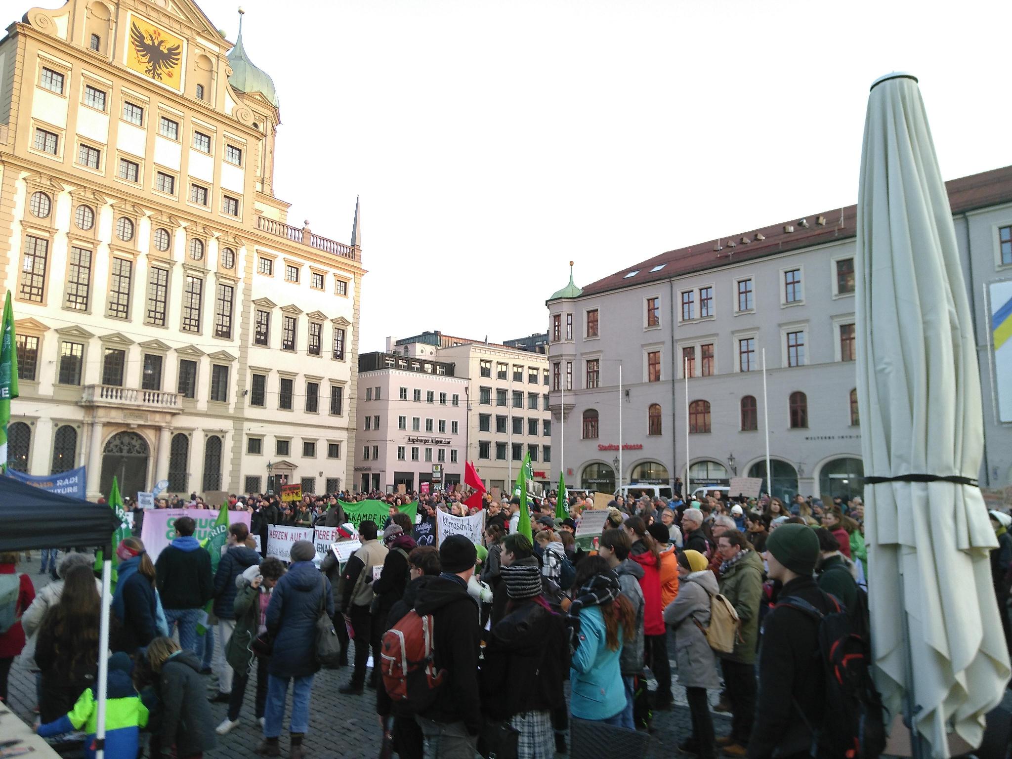 Für die Abschlussworte stehen hunderte Menschen im Halbkreis um eine Bühne auf dem Königsplatz. Links im Hintergrund thront das durch die Sonne angeleuchtete Rathaus.