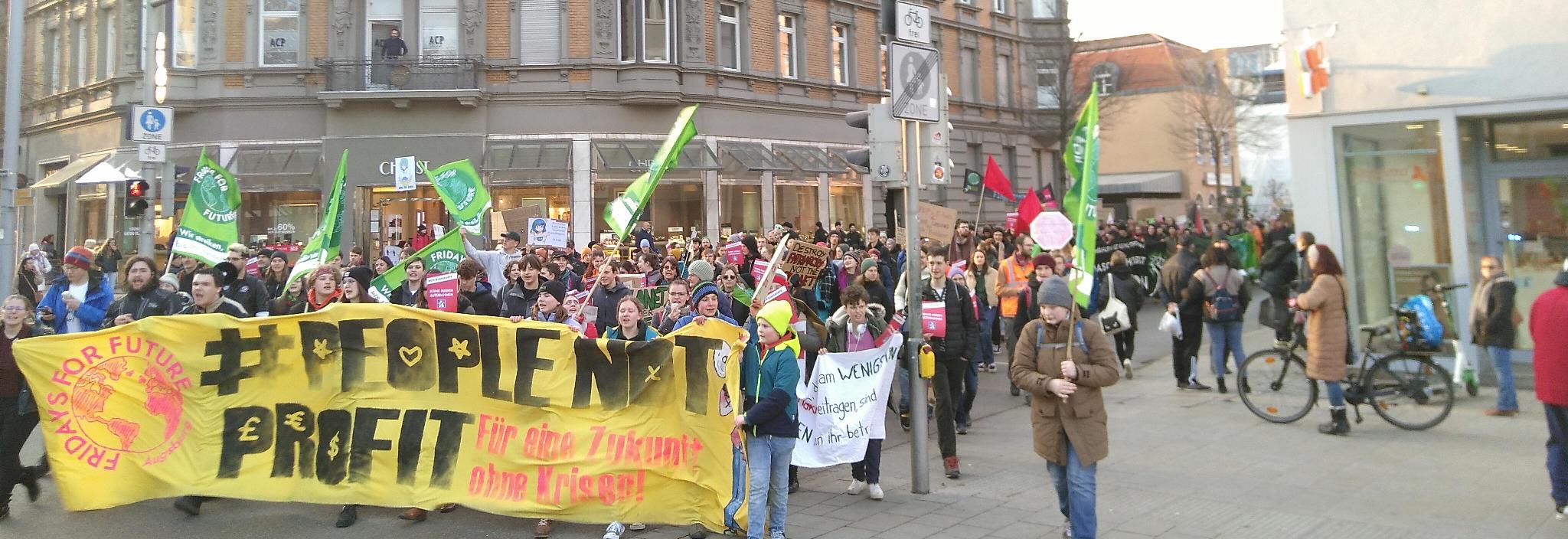 Die Spitze des Demonstrationszuges erreicht aus der Schaezlerstraße kommend den Rathausplatz. Vornweg laufen Kinder und junge Erwachsene, die ein breites Banner mit den Schriftzügen „FRIDAYS FOR FUTURE“, „#PEOPLE NOT PROFIT“ und „Für eine Zukunft ohne Krisen!“ vor sich her tragen. Dahinter läuft eine große Menge vorwiegend junger Menschen. Viele von ihnen tragen deutlich sichtbar Zettel mit der Aufschrift „KEINE NEUEN AUTOBAHNEN“. Mindestens ein halbes Dutzend ‚Fridays for Future‘-Fahnen werden mitgetragen. Auf einem Pappschild steht „DESTROY PATRIARCHY NOT THE PLANET“. Es gibt weitere Pappschilder und Banner in der Menge, deren Aufschrift auf dem Bild aber nicht zu entziffern ist. Einige dutzend Meter weiter hinten im Demozug sind noch einige rote Fahnen auszumachen. Dieser Bereich wird von einigen Polizist*innen flankiert.