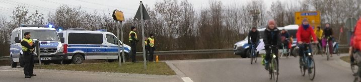 Eine Abzweigung nahe Friedberg: Drei Einsatzwägen bewachen eine Abzweigung in Richtung A8. Zwischen den Einsatzwägen stehen mindestens vier Polizist*innen und beäugen die an ihnen vorbei fahrende Fahrraddemo. In den Einsatzwägen sind weitere Polizist*innen zu erkennen.