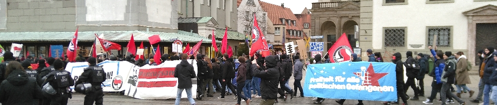 Eine antifaschistische Demo zieht vor dem Klimacamp vorbei. In einigem Abstand flankiert wird sie von einer großen Anzahl an Polizist*innen. In der Demo gibt es viele Fahnen mit der Aufschrift „Antifaschistische Aktion“. Auf Bannern an der Seite des dicht an dicht laufenden Demonstrationszuges stehen Sprüche wie „Freiheit für alle politischen Gefangenen“, „#unbeugsam“ und „Kampf der Klassenjustiz“.