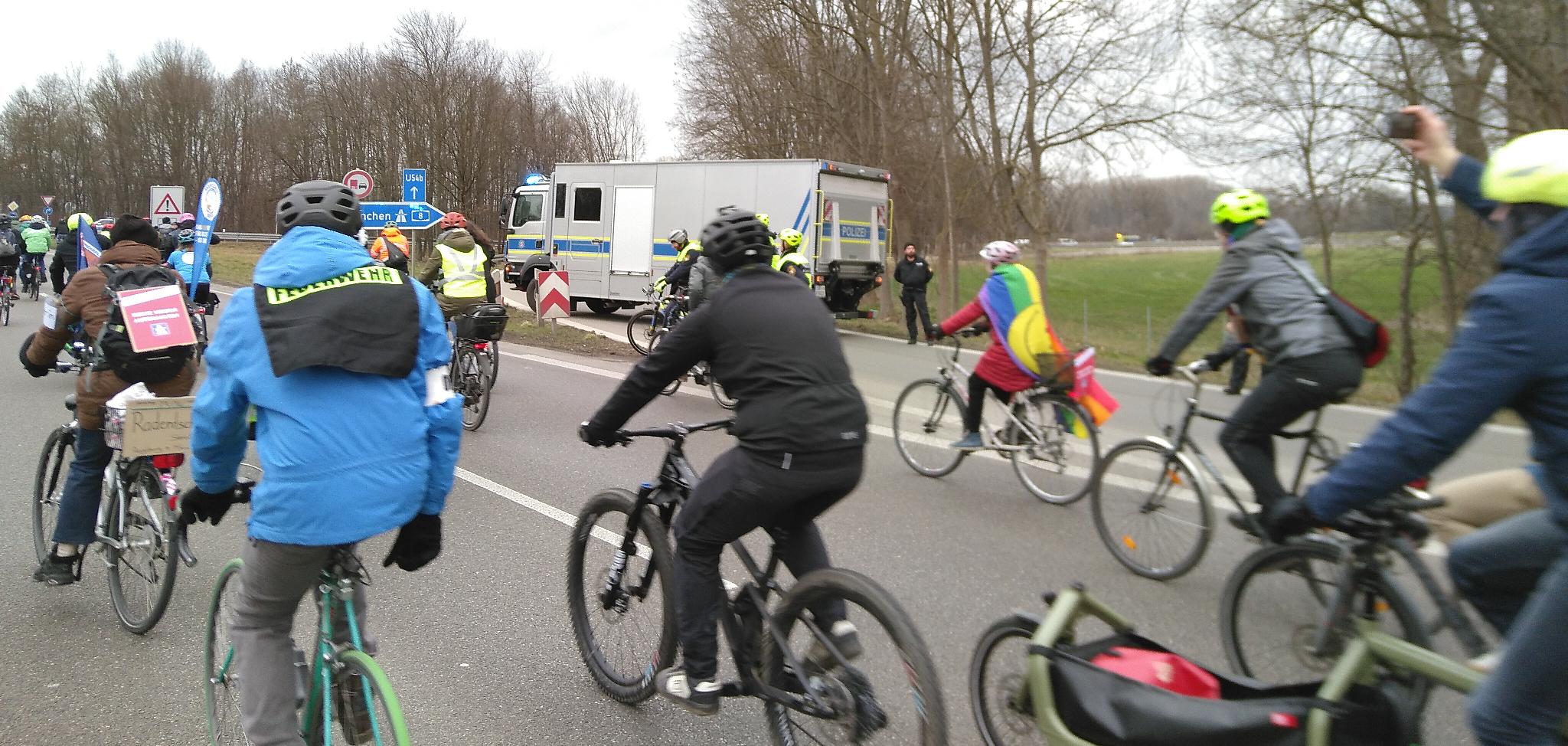 Eine bunte Gruppe an Fahrradfahrer*innen fährt der Auffahrt auf die A8 vorbei. Quer auf der Auffahrt steht ein großes lastwagenartiges Fahrzeug der Polizei. Davor befinden sich Polizist*innen, zum Teil auf Fahrrädern. Rechts neben dem Fahrzeug sieht man im Hintergrund zwischen einigen Bäumen die Autobahn und vereinzelte Fahrzeuge.