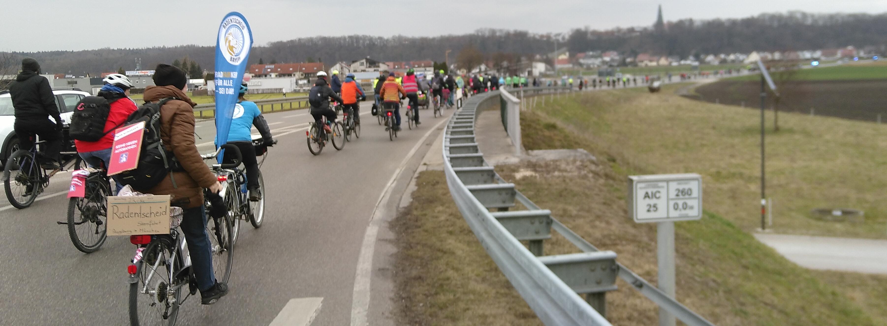 Demonstrationsteilnehmer*innen fahren nördlich der A8 auf einer Straße nahe Derching. Man erkennt eine Fahne, die auf den bayerischen Radentscheid wirbt, Pappschilder, welches auf eine anstehende Sternfahrt nach München verweist, und ein „KEINE NEUEN AUTOBAHNEN“-Schild.