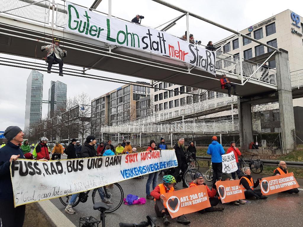 Die Demonstration posiert mit Bannern: Auf der Fußgängerbrücke stehen Menschen mit Kameras und Megafon. Am Brückengeländer hängt ein Banner mit der Aufschrift „Guter Lohn statt neue Straßen!“. Links und rechts des Banners hängt jeweils ein*e Aktivist*in an Seilen von der Brücke. Mehrere Meter tiefer auf der Autobahn stehen zahlreiche Fahrraddemonstrant*innen hinter weiteren Bannern. Diese sind beschriftet mit „FUSS, RAD, BUS UND BAHN STATT AUTOWAHN“, „1,5 Grad, neun Tote und 1053 Verletzte pro Tag“, „VERKEHRSWENDE JETZT“, „verkehrswende.siehe.website“, „ART. 20A GG = LEBEN SCHÜTZEN“ und „LETZTE GENERATION VOR DEN KIPPPUNKTEN“.