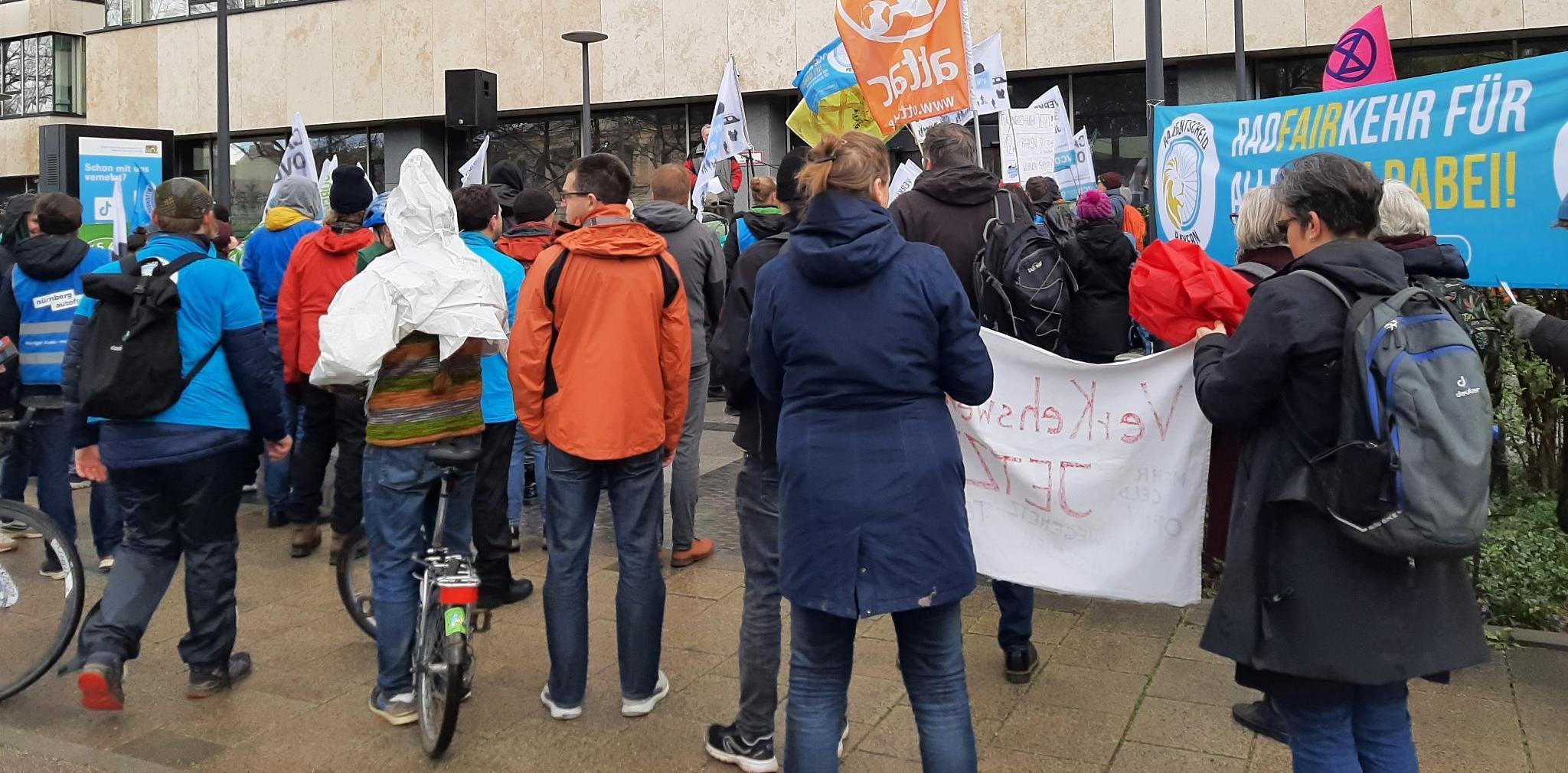 Mehrere Menschen stehen vor einem Podium, welches vor bayerischen Staatsministerium für Wohnen, Bau & Verkehr aufgebaut ist. Es gibt viele Fahnen und Banner, darunter von „Radentscheid Bayern“ und „attac“.