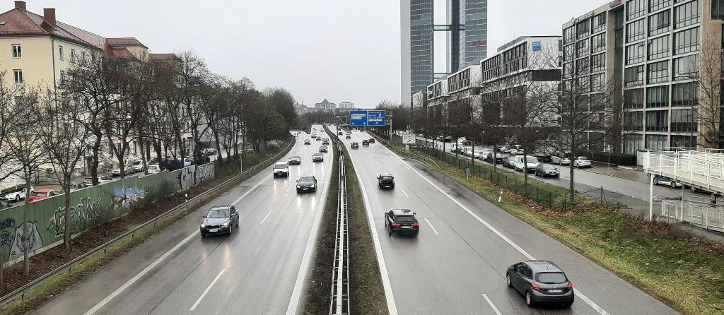 Das Bild wurde von der Brücke über die Autobahn aufgenommen. Es zeigt den entsprechenden Autobahnabschnitt. Links und rechts ist die Autobahn von Wohn- und Bürohäusern eingesäumt. 