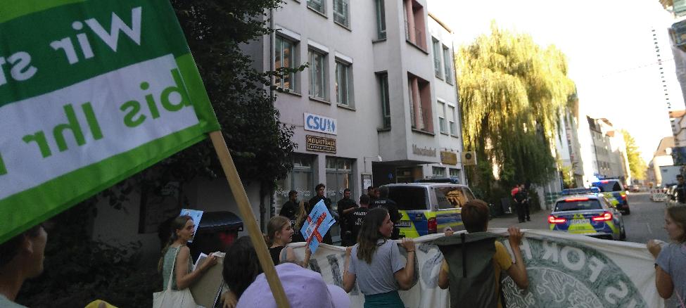 Ein Polizeieinsatzwagen und mehrere Polizist*innen stehen vor dem CSU-Büro. Im Hintergrund am Gebäude hängt ein CSU-Logo. Links neben dem Logo im Bild eine grüne „Wir streiken, bis ihr handelt!“-Fahne. Ein Einsatzwagen und ein Streifenwagen fahren vor der Demo her. Mehrere weitere Polizist*innen laufen neben der Spitze der Demo. 