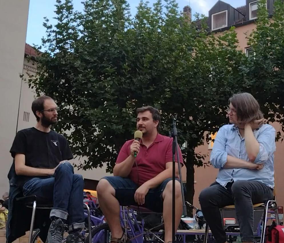 Dei Mensche nsitzen auf Stühlen auf einem Podium am Moritzplatz. Die mittlere Person hält ein Mikrofon in der Hand. Die beiden Personen an der Seite lauschen.