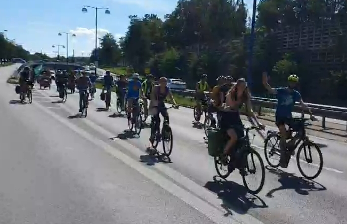 Die Fahrraddemo fährt über die B17. Einige Aktivist*innen auf Fahrrädern winken in Richtung der Kamera. Im Hintergrund sind vereinzelte Fahrzeuge auf der durch einen Grünstreifen und Leitplanken abgetrennten Gegenfahrbahn.