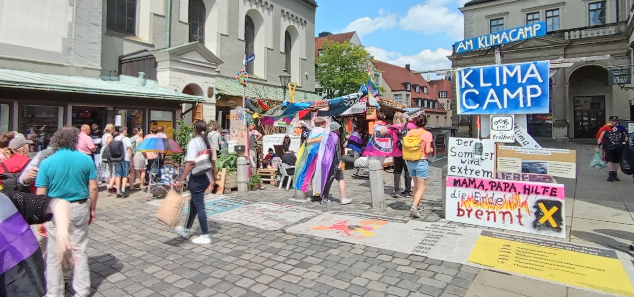 Foto des Klimacamps mit Regenbogenmfahnen und buntem Treiben in Pridefarben.