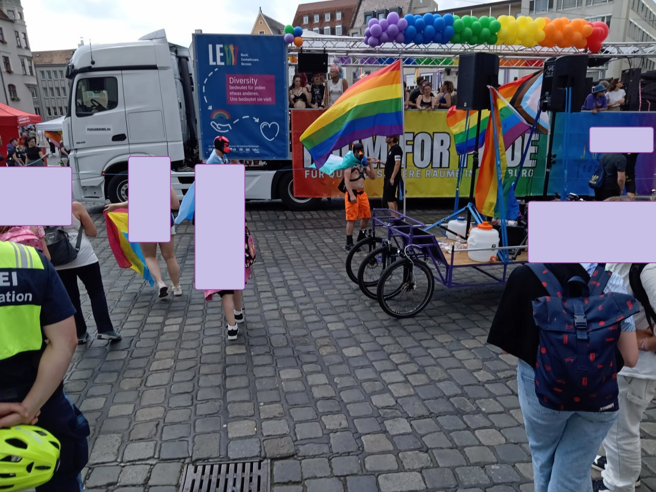 Foto eines Party-Lasters mit Luftballons in Farben der Regenbogenflagge und feiernder Menge ab Bord. Im Vordergrund das Sechsrädrige Lastenrad vom Klimacamp.