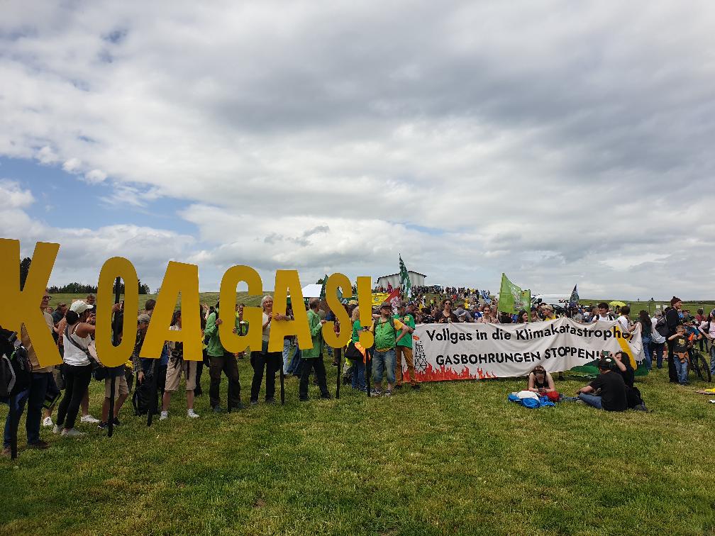 Auf einer Wiese in Handlage stehen hunderte Menschen. Vornen links halten Menschen die personengroßen Buchstaben „KOA GAS!“ in die Höhe. Daneben steht das Banner mit der Aufschrift „Vollgas in die Klimakatastrophe?! GASBOHRUNGEN STOPPEN!“. Die Menschen stehen und sitzen auf der Wiese, lachen und unterhalten sich. 