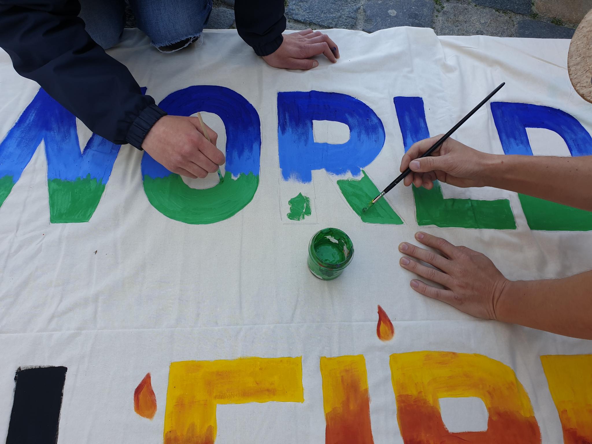 Ein Banner liegt auf Kopfsteinpflasterboden. Menschen knien darauf und malen mit Pinseln. Die Wörter „WORLD“ und „FIRE“ sind lesbar.