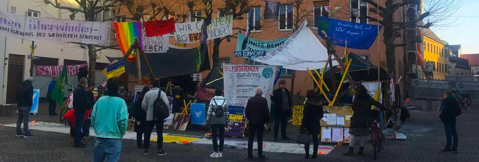 Unser Klimacamp am Moritzplatz zu Jahresbeginn 2022: Vor dem Camp haben sich etwa ein dutzend Interessierte versammelt und lesen die zahlreichen Banner und Plakete. Ein Vertreter des Camps erzählt von der Arbeit des Camps.